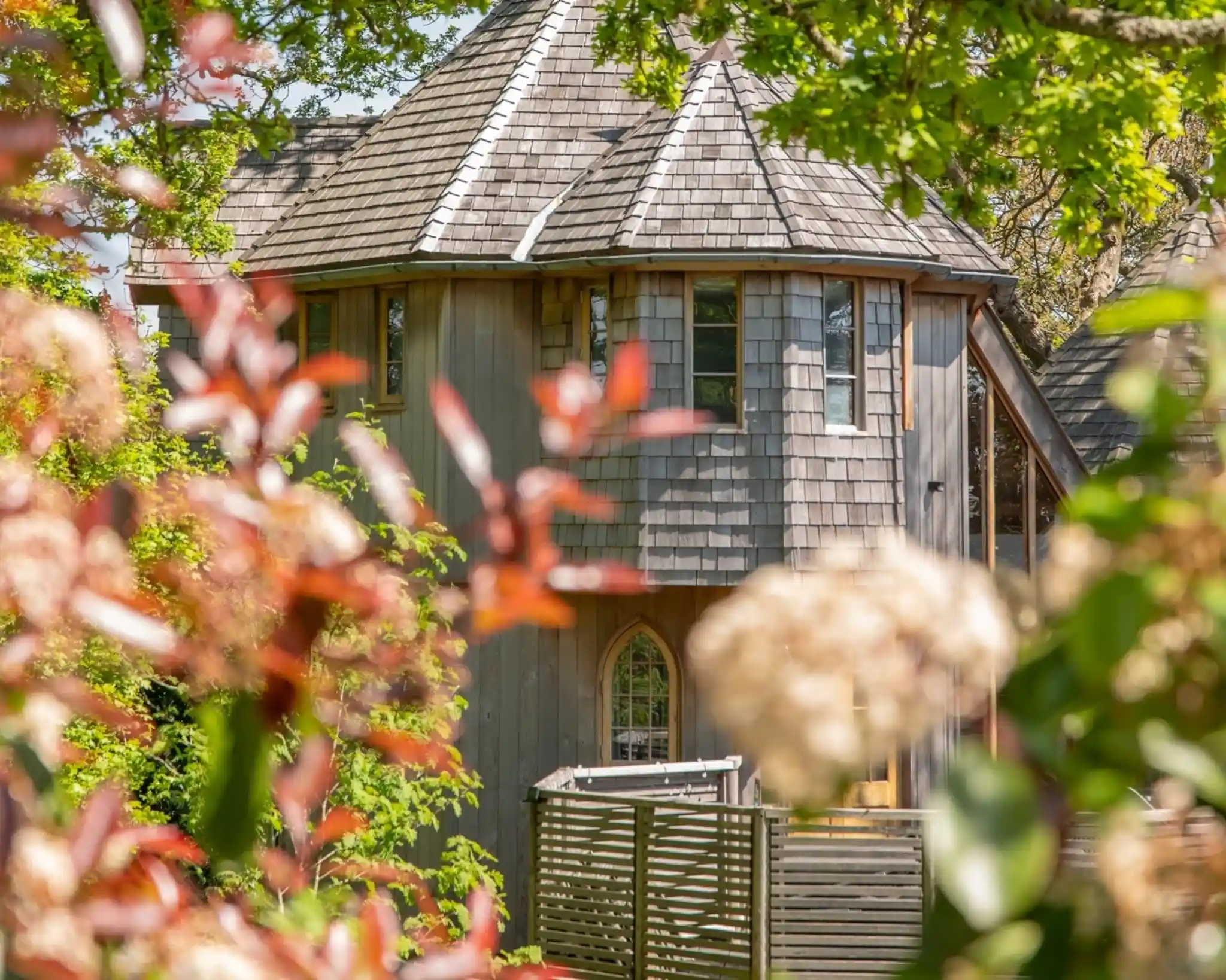 A charming wooden house with a unique turret design, surrounded by lush greenery and vibrant foliage. The structure features a shingled roof and large windows, blending harmoniously with its natural setting.