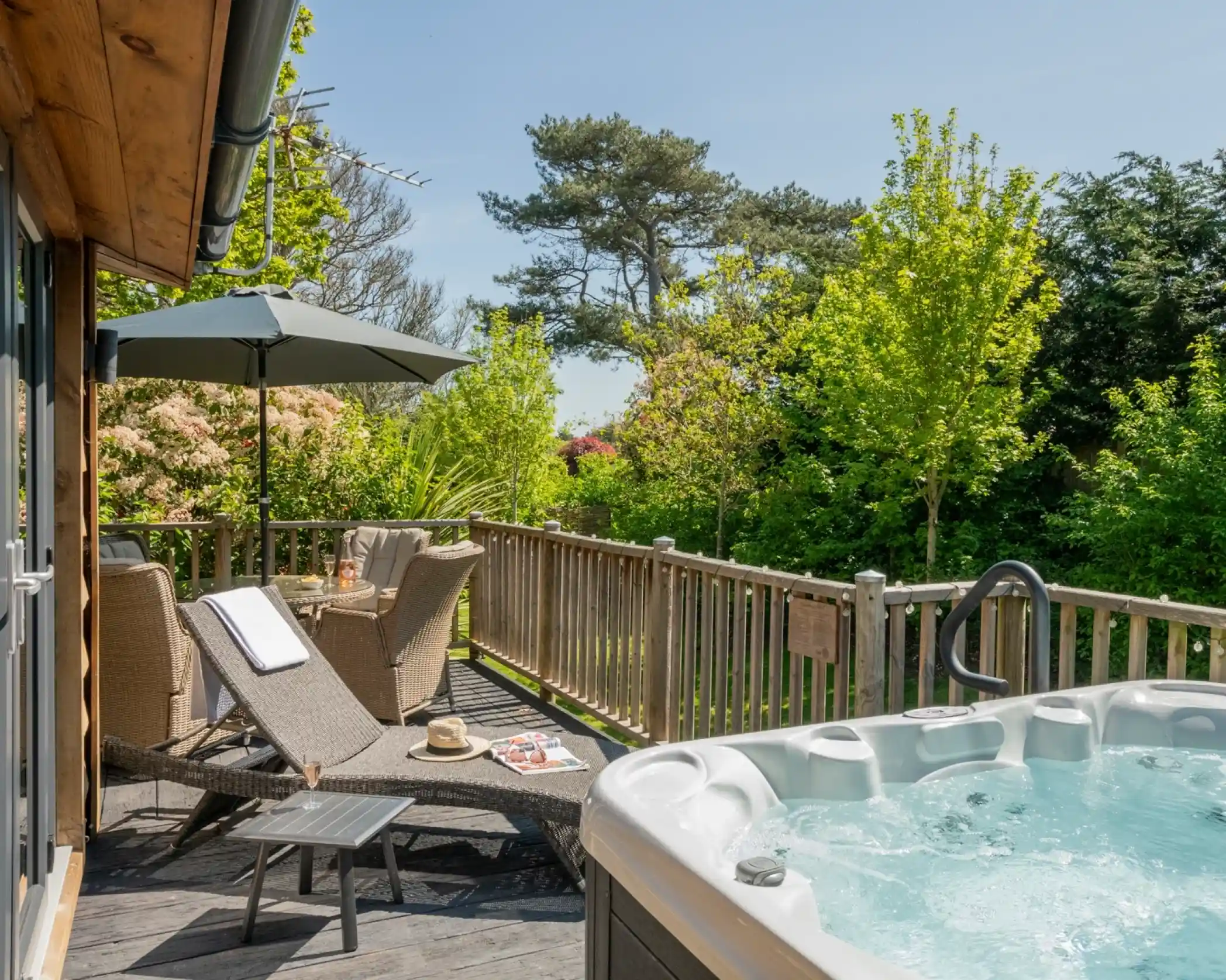 A wooden deck features a hot tub, two lounge chairs, and an umbrella. Lush greenery surrounds the area, creating a serene outdoor space.