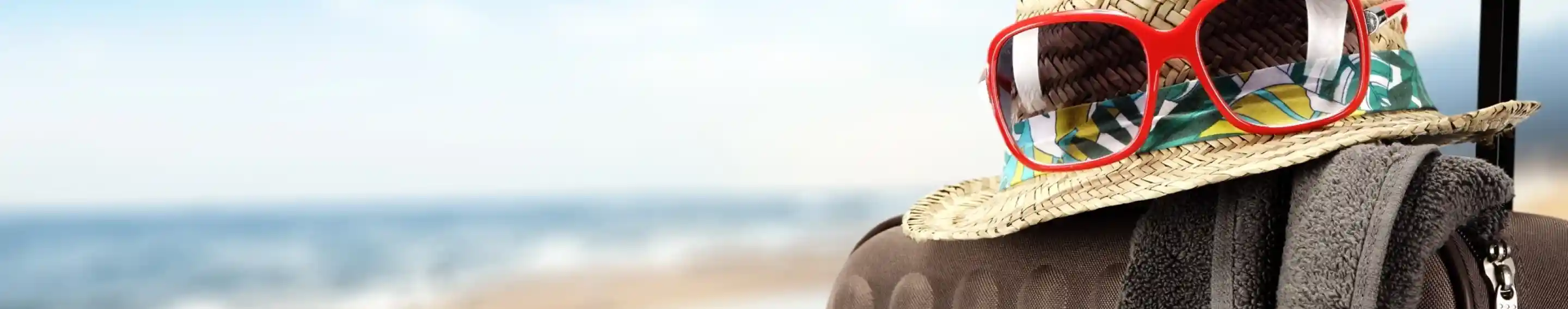 A straw hat with red sunglasses rests on a suitcase, accompanied by a beach towel, with a blurred seaside background.