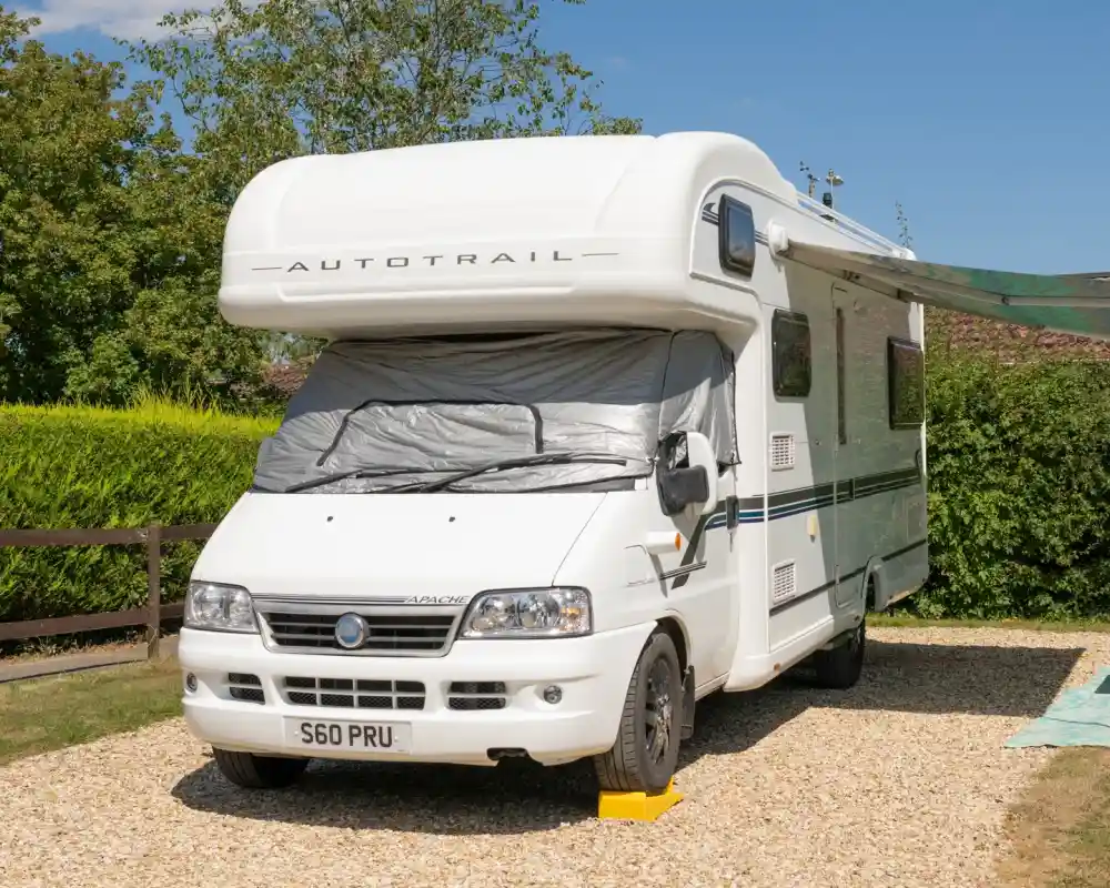 A white motorhome parked on a gravel area, partially covered with a silver sunshade. Green foliage and a clear blue sky are in the background.
