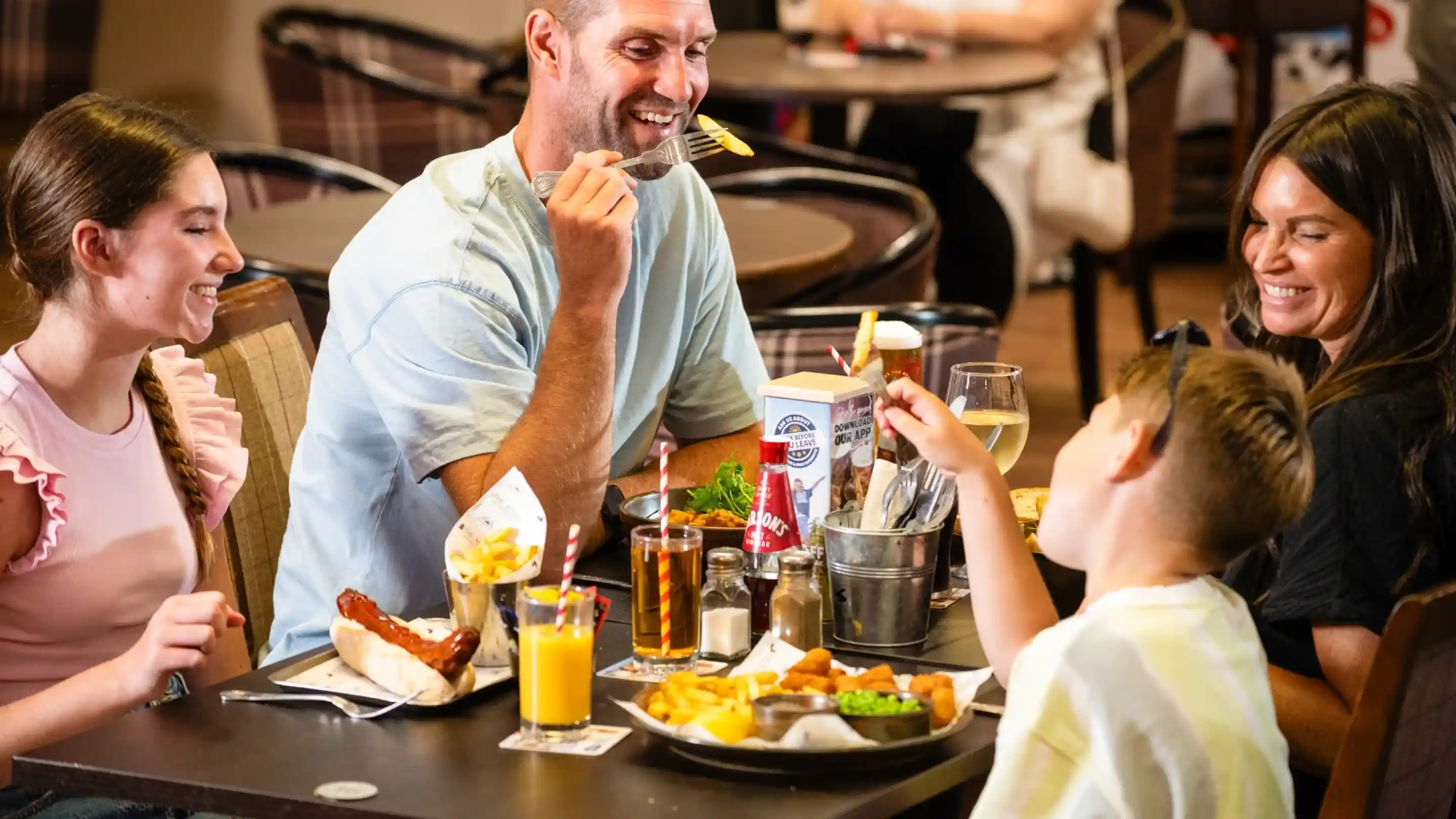 A family enjoying a meal together in a casual dining setting. They are seated at a table with various dishes, including hot dogs, fries, and drinks. The group consists of four people, with a child playfully pointing at one of the adults. Laughter and smiles are evident among them.