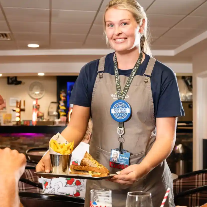A smiling waitress serves a tray with a sandwich and fries to a couple at a restaurant table. The woman looks on while the man engages with the waitress. Decor and bar can be seen in the background.