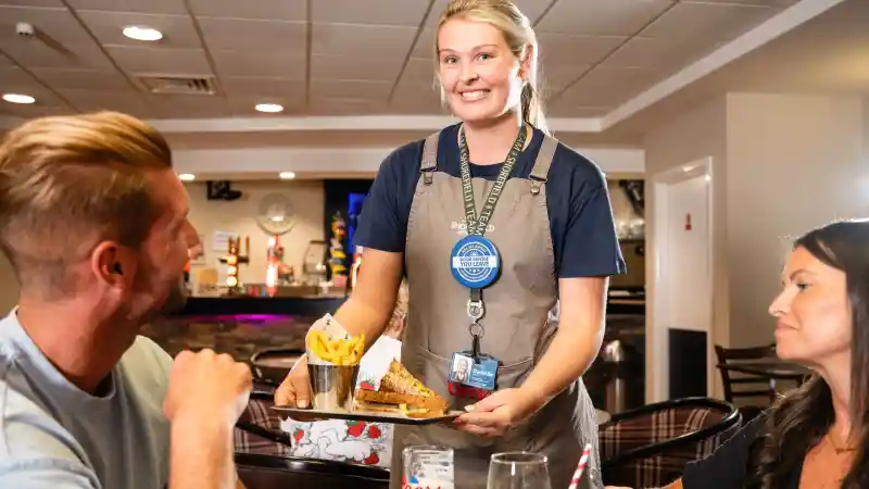 A smiling waitress serves a tray with a sandwich and fries to a couple at a restaurant table. The woman looks on while the man engages with the waitress. Decor and bar can be seen in the background.