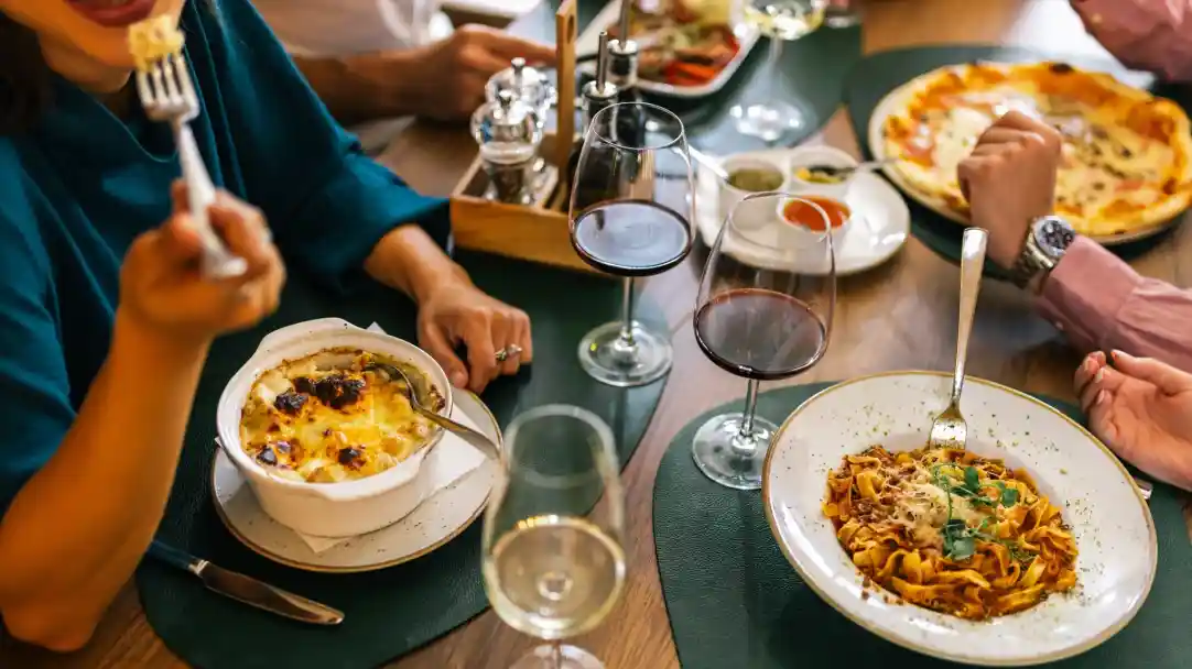 A table set with various dishes, including a bowl of baked pasta, a plate of spaghetti, and a pizza. Several glasses of red wine are present, along with condiments and a wooden serving tray. A person is holding a fork with food in the foreground.