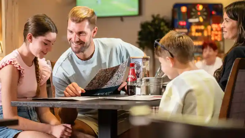 A man smiles while showing a menu to a girl and a boy at a table in a casual dining setting. The girl has long hair and wears a pink top, while the boy is wearing sunglasses. A woman sits nearby, watching them. The background features a sports screen and a vending machine.