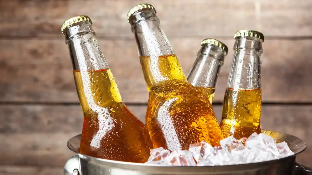 Several glass bottles of beer are submerged in ice in a metal bucket. The bottles have condensation on the surface, and the golden liquid inside is illuminated against a rustic wooden background.