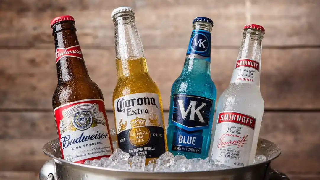 A variety of beer and flavored alcoholic beverages in glass bottles, displayed in a chilled ice bucket. The brands include Budweiser, Corona Extra, VK Blue, and Smirnoff Ice.