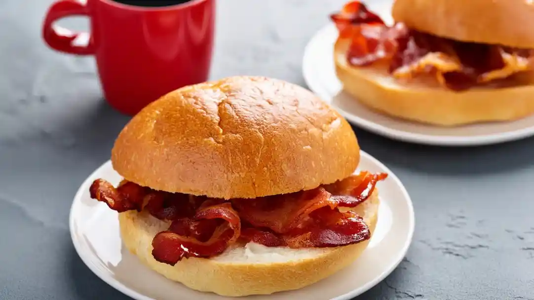 Two plates with freshly made bacon sandwiches on a textured surface. The sandwiches consist of a soft, golden-brown bun filled with crispy bacon. A small red cup with dark liquid is in the background.