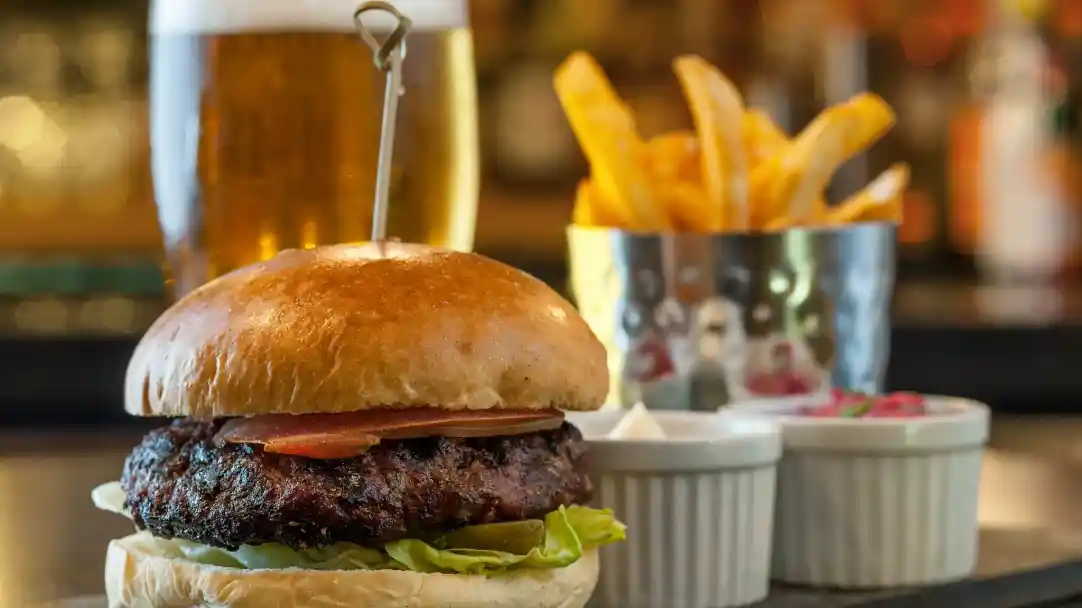 A freshly made burger with a toasted bun, lettuce, and tomato sits in the foreground, accompanied by small bowls of sauces. Behind it, a glass of golden beer and a metallic cup filled with crispy French fries are visible.