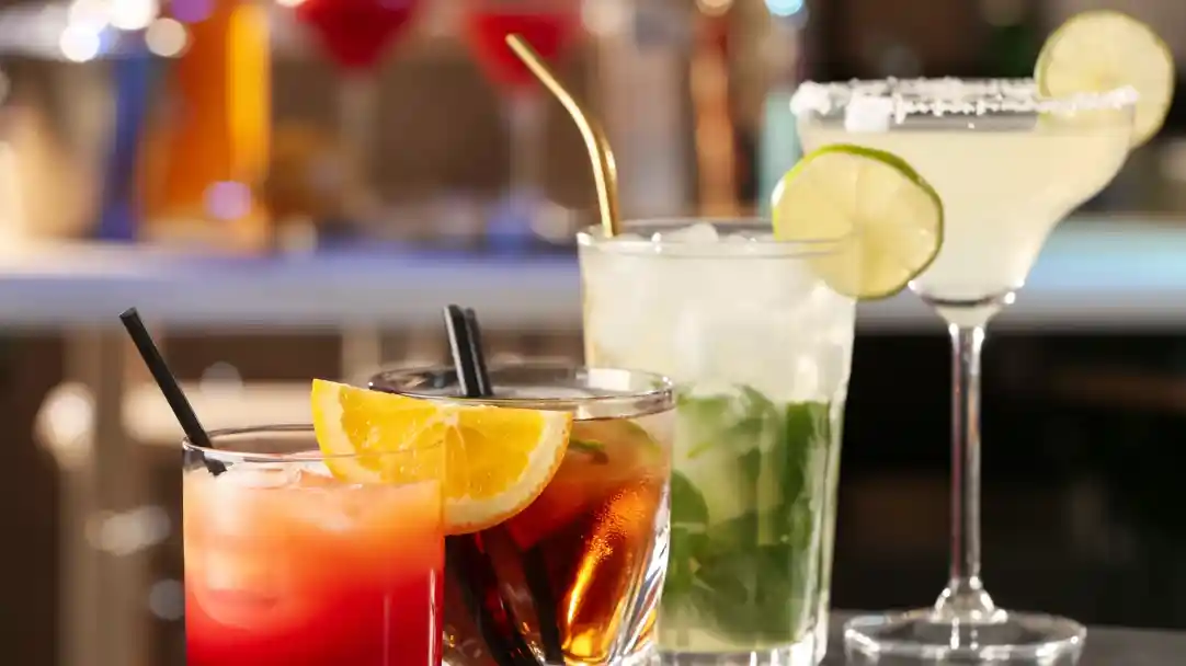 A close-up of four colorful cocktails on a bar. From left to right: a fruity red drink garnished with orange slices, a dark drink with a straw, a refreshing mojito with mint, and a margarita with a salted rim and lime wedge.