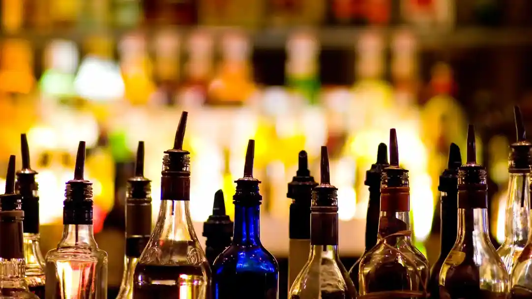 Various liquor bottles with distinct shapes and colors are lined up, with a blurred bar background showcasing colorful backlit liquids.