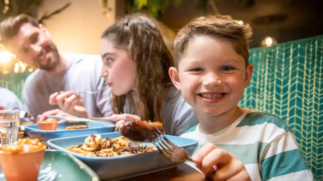 A young boy smiles while holding a fork with food on it at a dining table. A girl beside him is eating, and a man in the background is looking at them. The table is set with plates of food and drinks.