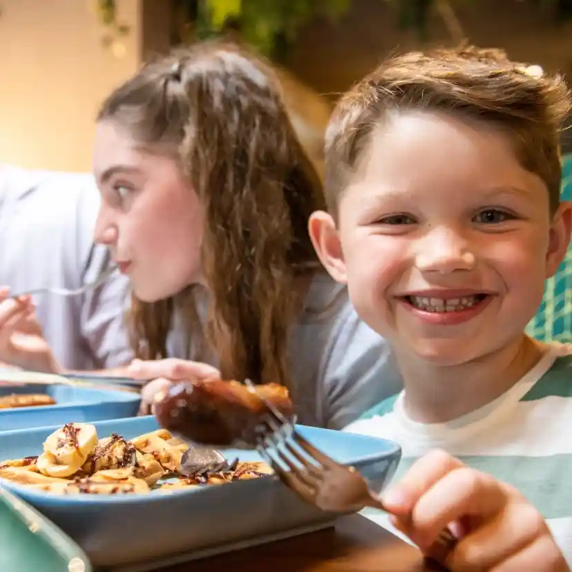 A young boy smiles while holding a fork with food on it at a dining table. A girl beside him is eating, and a man in the background is looking at them. The table is set with plates of food and drinks.
