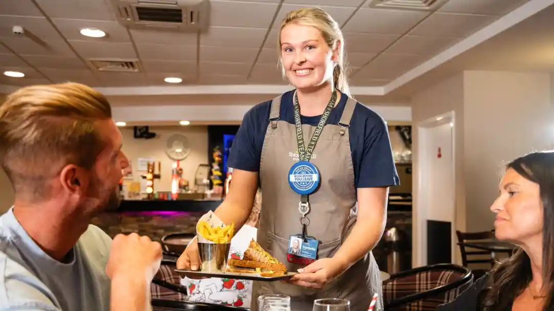 A waitress smiles while serving a plate with food and fries to two seated customers at a restaurant. The setting is casual, with a background featuring bar equipment.