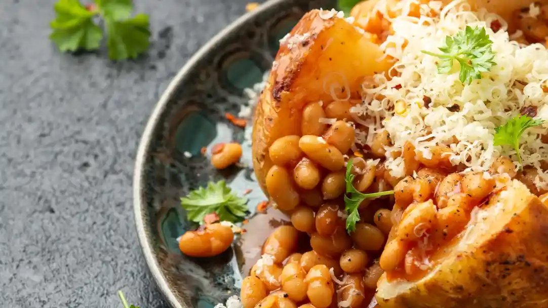 A close-up of a plate featuring baked potatoes filled with baked beans, topped with shredded cheese and garnished with fresh herbs. The dish is presented on a decorative blue and green plate.