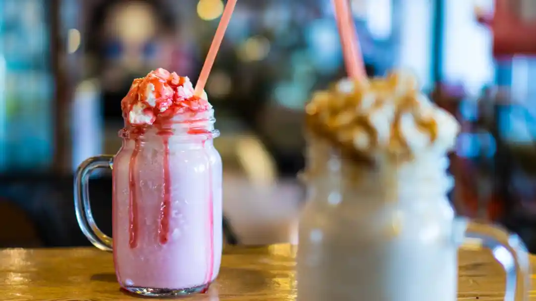Two mason jars filled with creamy milkshakes on a wooden table. One milkshake is pink with whipped cream and strawberry syrup, while the other is beige with caramel drizzle and whipped cream. Soft blurred background.