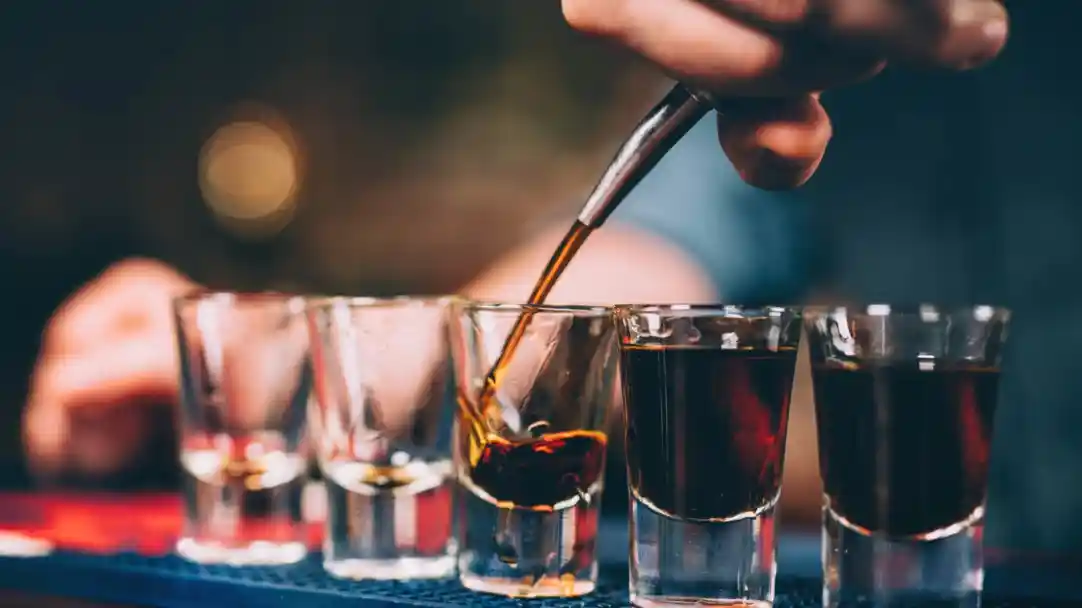 A hand pours a dark liquid into a shot glass, part of a lineup of several shot glasses on a bar counter. The glasses vary in contents, with some filled with clear liquid and others with darker drinks.