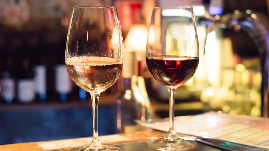 Two wine glasses sit on a bar counter, one filled with white wine and the other with red wine. The background features soft lighting and blurred bottles.