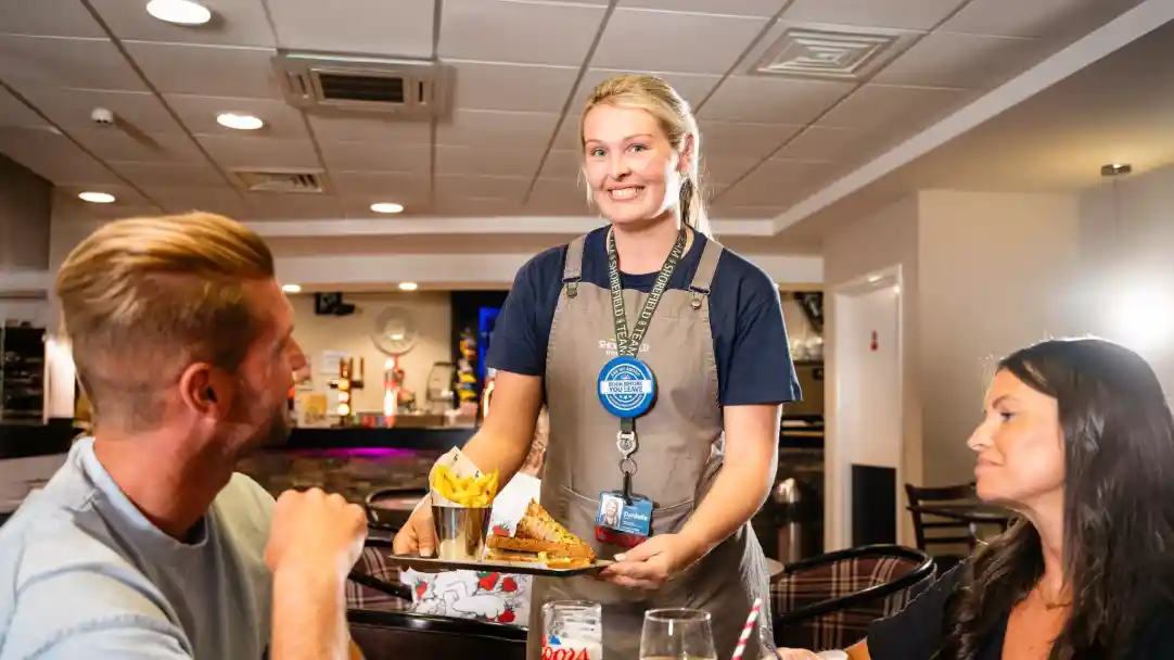 A waitress in an apron smiles while serving a plate of food to two customers at a restaurant. The interior features a bar area and casual dining.