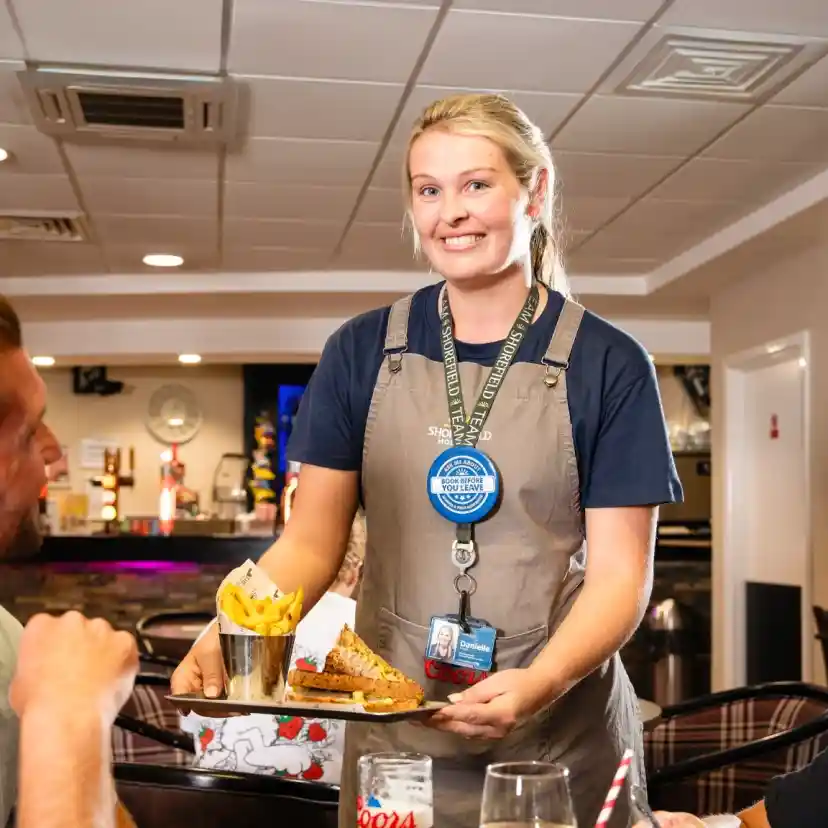 A waitress in an apron smiles while serving a plate of food to two customers at a restaurant. The interior features a bar area and casual dining.