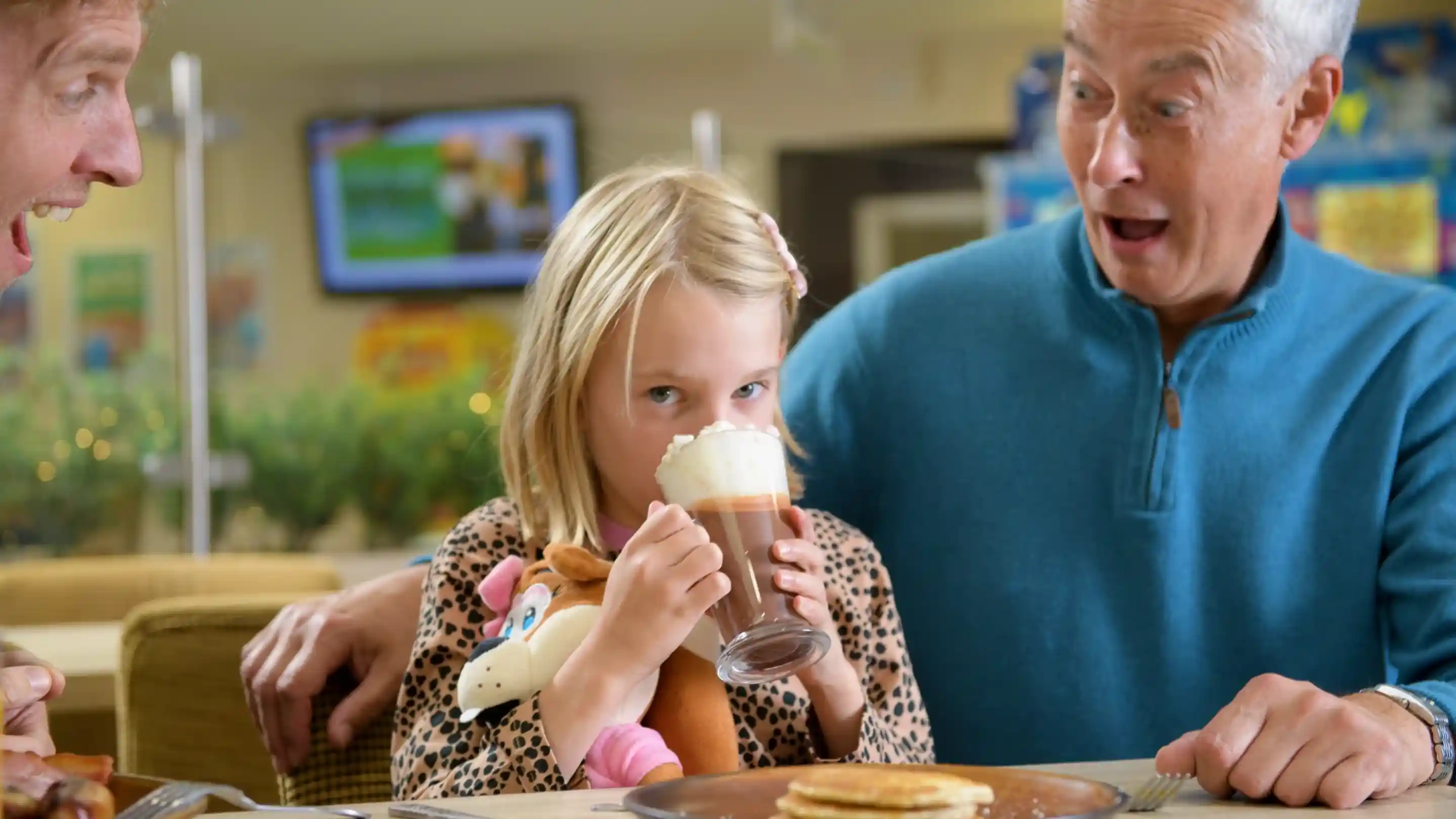 A young girl with a short blonde bob holds a milkshake while sitting at a table. She has a stuffed toy horse beside her and a playful expression as she looks at two adults, one of whom is surprised. Pancakes are on the table in front of them.