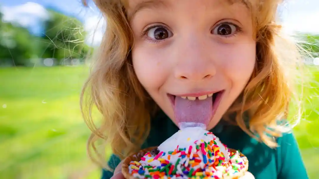 A joyful child with curly hair relishes an ice cream cone topped with colorful sprinkles, sticking out their tongue in excitement. A grassy landscape is visible in the background.