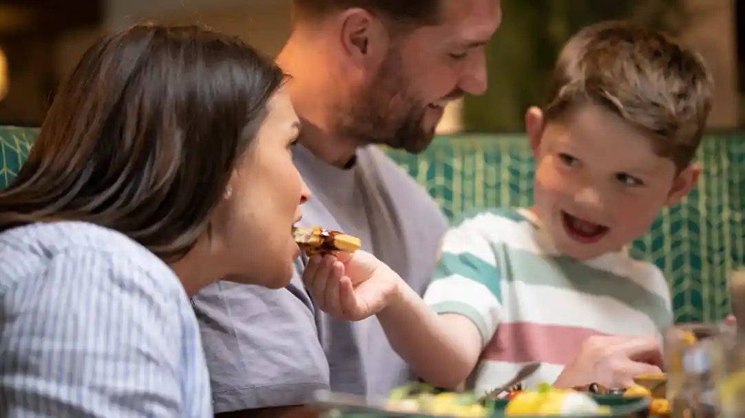 A woman is taking a bite of food while a smiling young boy feeds her another piece. A man sits beside them, watching with a smile, in a cozy dining setting.