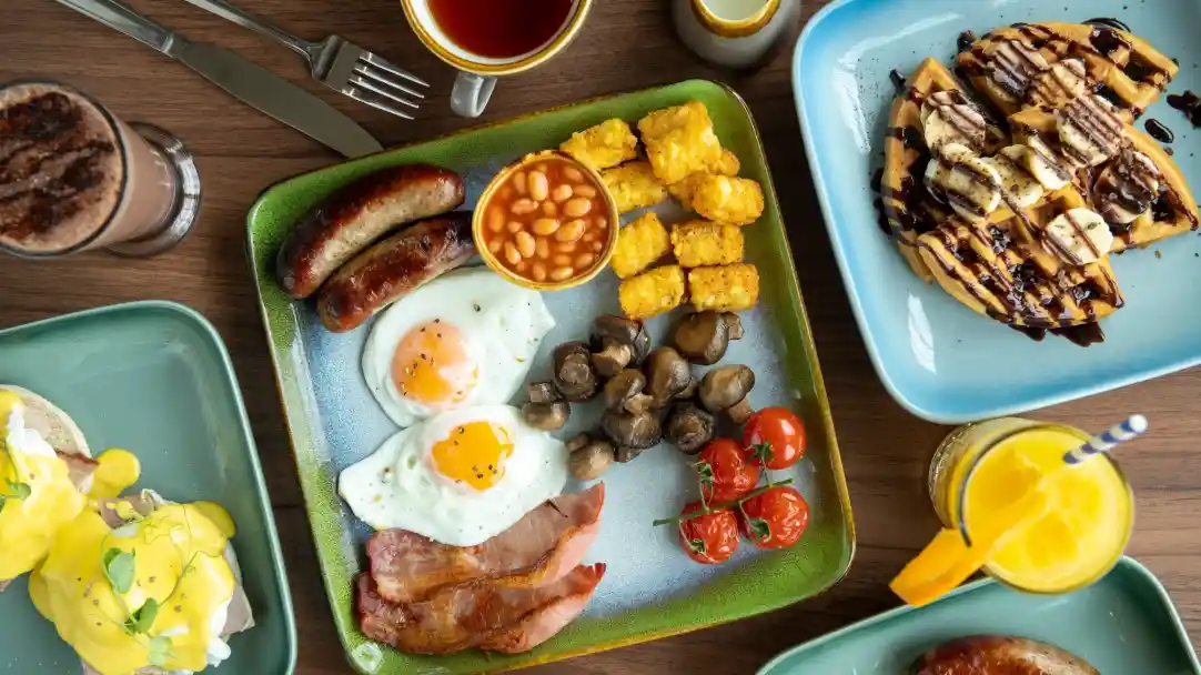 A colorful breakfast spread featuring a plate with two fried eggs, bacon, sausages, baked beans, sautéed mushrooms, and cherry tomatoes. Nearby, a serving of golden hash browns and a plate of waffles drizzled with chocolate sauce. Beverages include coffee and a tropical drink with a straw.
