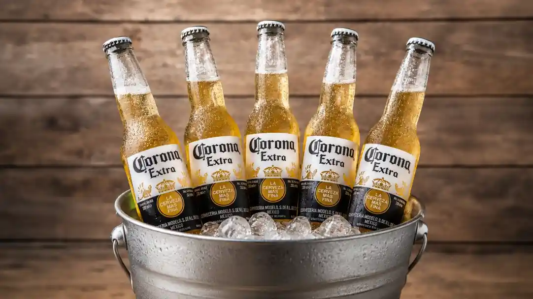 Five bottles of Corona Extra beer are nestled in a bucket filled with ice. The background features rustic wooden boards that enhance the chilly vibe.