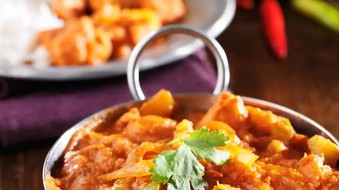 A close-up of a serving dish filled with a vibrant orange curry topped with fresh cilantro leaves. In the background, another dish of food is slightly out of focus. The setting includes a dark wooden surface and hints of red chilies.
