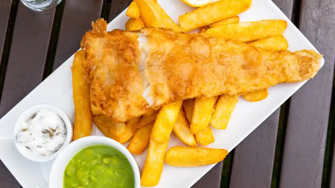A serving of crispy fried fish with golden French fries, accompanied by a wedge of lemon and two dipping sauces—one tartar and one pea puree.