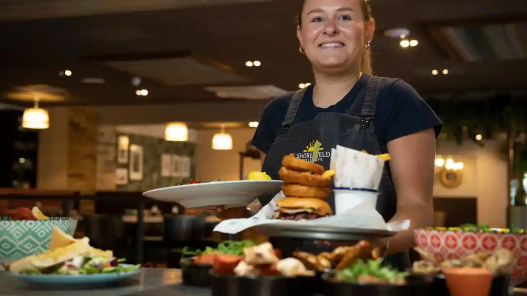 A smiling server wearing an apron holds two plates, one stacked with onion rings and the other with salad. In the background, a cozy restaurant atmosphere is visible, with various dishes and greenery on the table.