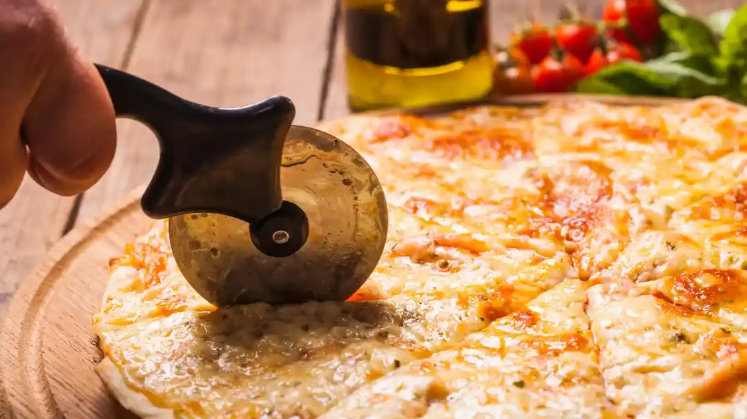 A hand using a pizza cutter to slice a cheese pizza on a wooden board, with a bottle of olive oil and fresh tomatoes in the background.