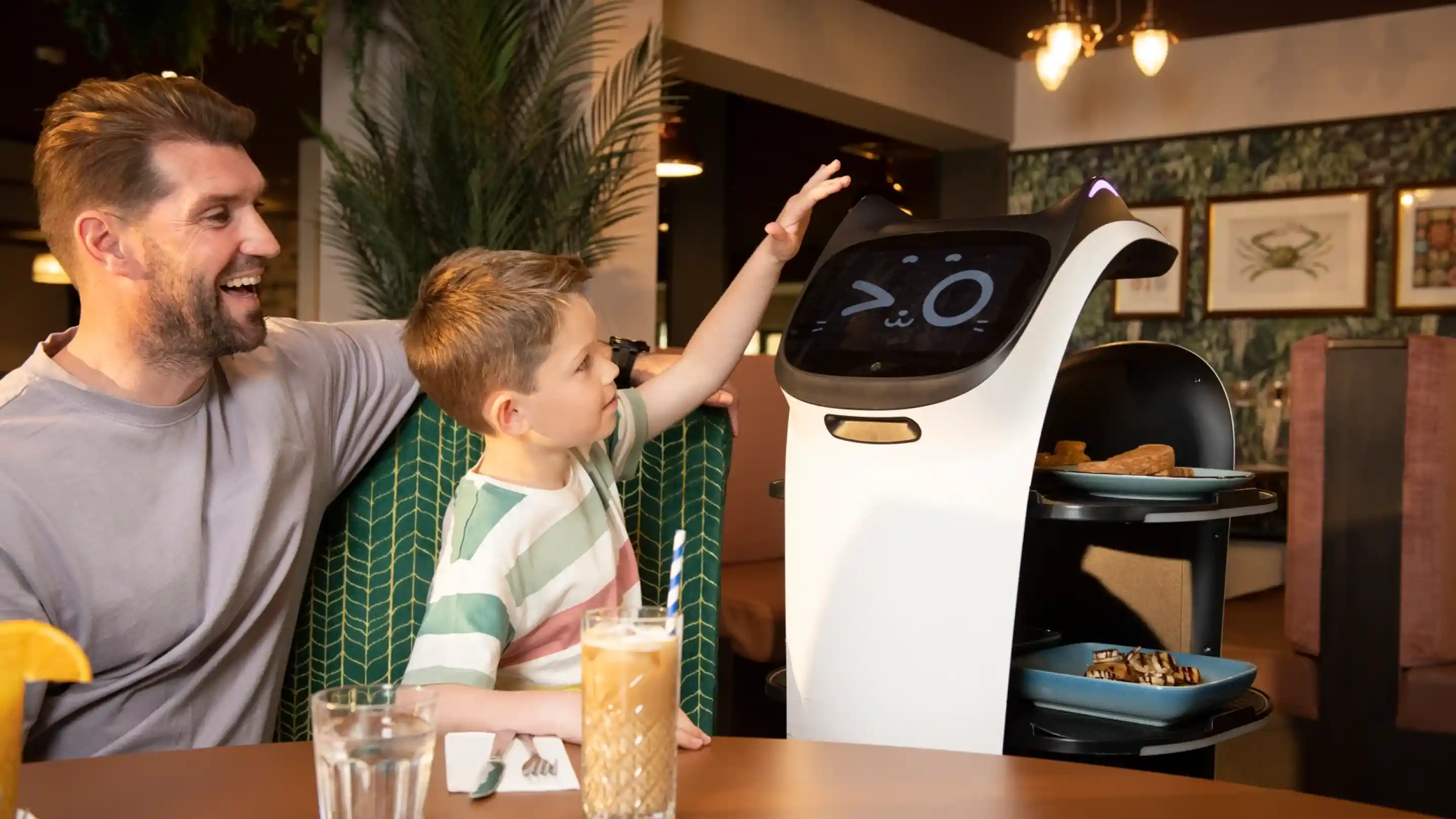 A father and son interact with a friendly-looking, humanoid robot in a restaurant. The child is reaching out to touch the robot's screen, which displays a cute face, while the father smiles beside him. The table is set with food and drinks, and the background features tropical decor.