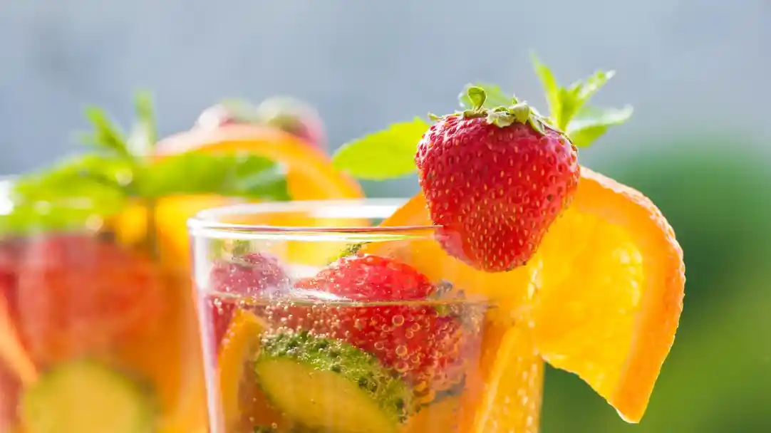 Sparkling beverage garnished with fresh strawberries, orange slices, and mint leaves, served in a clear glass against a blurred background.