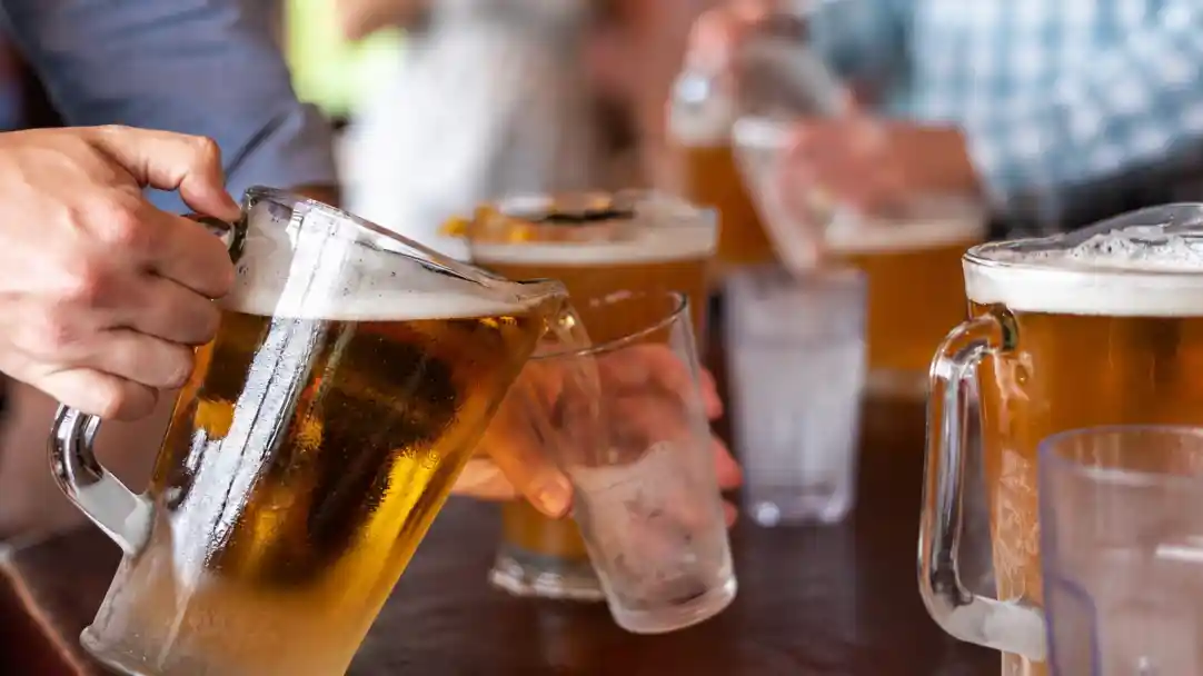 A person pours beer from a large pitcher into a glass, with several other glasses of beer visible in the background. The setting appears to be a social gathering or bar.