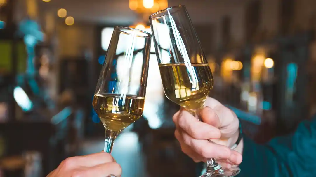 Two glasses of sparkling wine are clinking together in a toast, with a softly blurred warm, lit background suggesting a celebratory atmosphere.