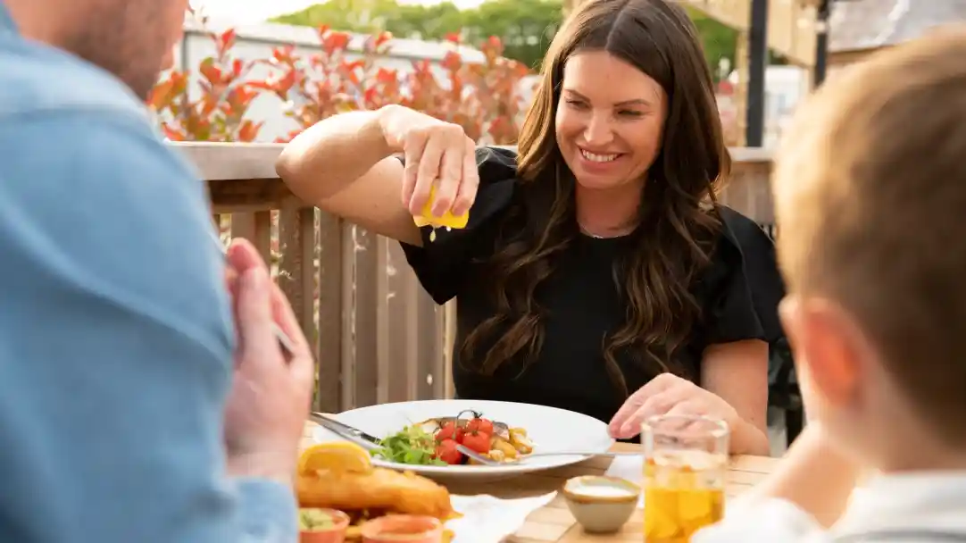 A woman with long, wavy brown hair is smiling as she squeezes lemon onto a plate of food while seated at a wooden table. In the foreground, a blurred figure is seen with hands clasped in a gesture of gratitude. The table is set with various dishes, and there are drinks nearby, along with colorful foliage in the background.