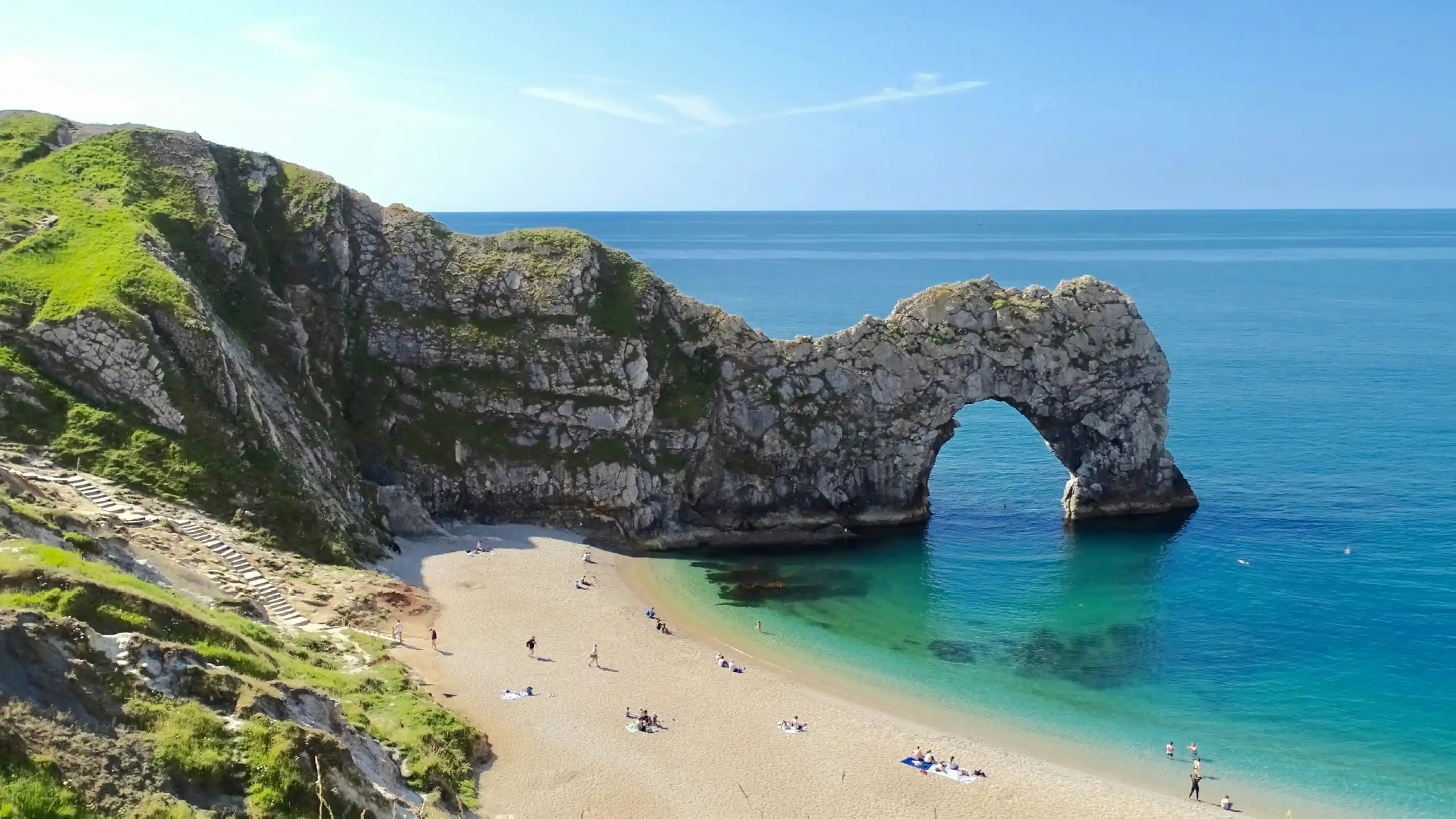 A stunning coastal view featuring the iconic Durdle Door rock formation rising from a sandy beach. Crystal-clear turquoise waters gently lap against the shore, while green cliffs provide a lush backdrop under a clear blue sky. Small groups of people are seen relaxing on the beach.