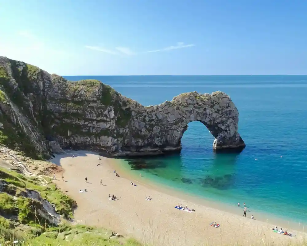 A scenic beach with golden sand and crystal-clear turquoise waters, featuring a large natural stone arch in the background. Lush green cliffs surround the area, and a few people are scattered across the beach enjoying the sunny weather.