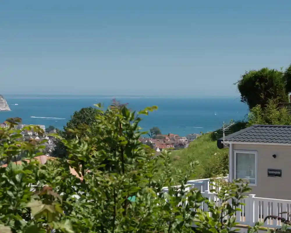 A serene coastal view featuring a blue ocean, small sailboats, and a distant shoreline. Lush greenery and a modest house with a sign partially visible in the foreground. Clear blue sky above enhances the picturesque scene.