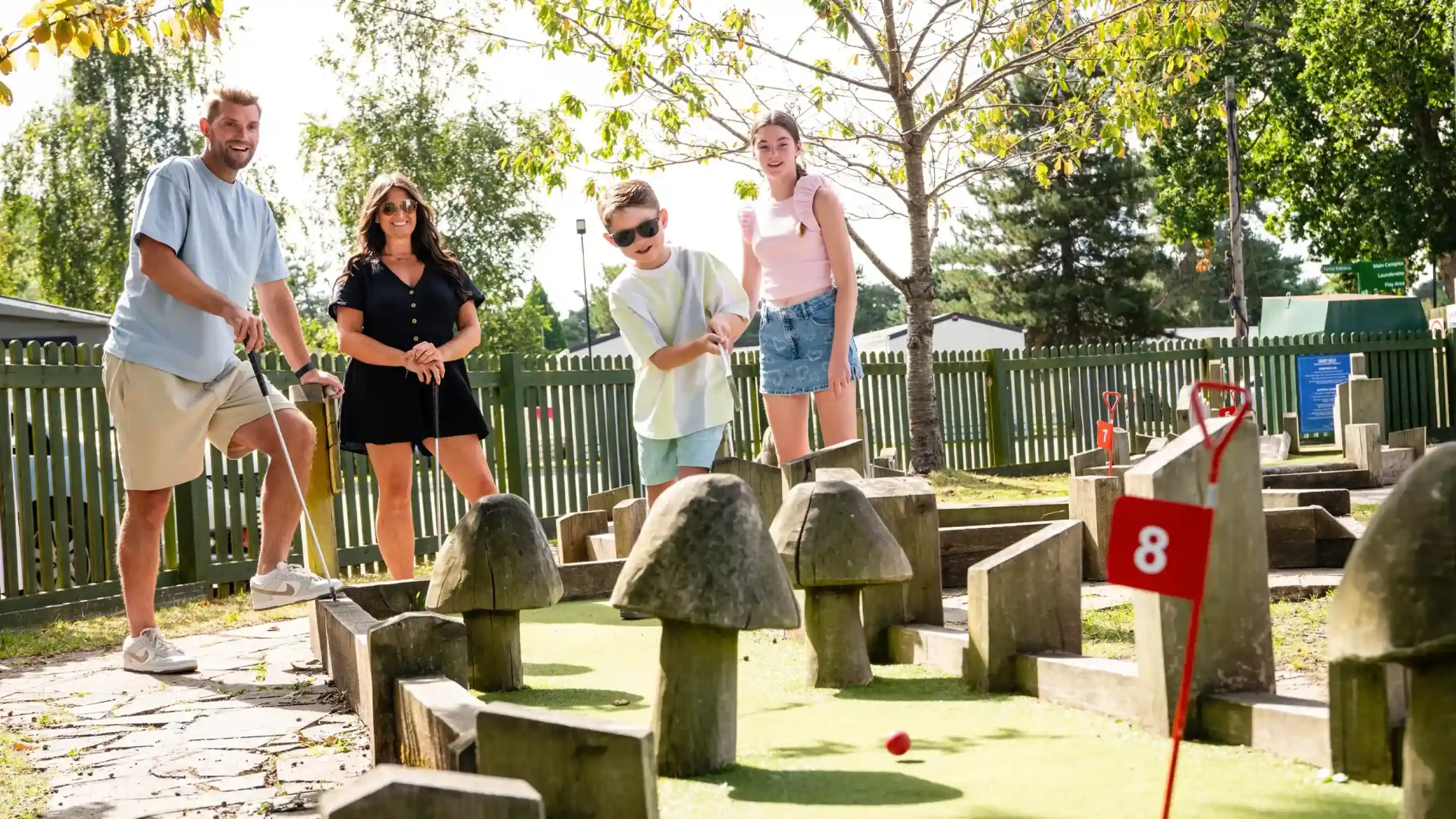 A family enjoys a game of mini-golf outdoors. A boy in sunglasses takes a shot while a woman and two others watch and cheer nearby. Surrounding the course are tall trees and a green fence.