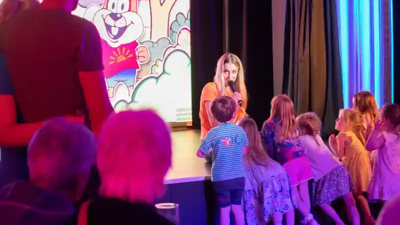 A young girl with long hair speaks into a microphone on stage, engaging a group of children gathered around her. A colorful animated screen featuring a cartoon character is in the background. Adults can be seen sitting in the foreground.