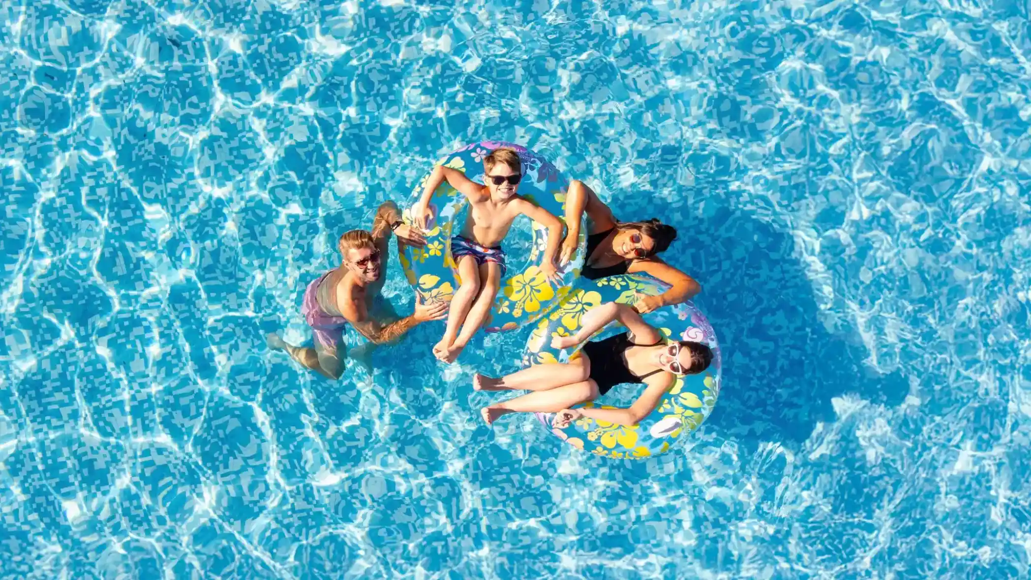 Aerial view of four people relaxing on colorful pool floats in a blue swimming pool. Two individuals are in the water, while two are lying on the floats, enjoying the sun. The pool's surface reflects light patterns creating a lively atmosphere.