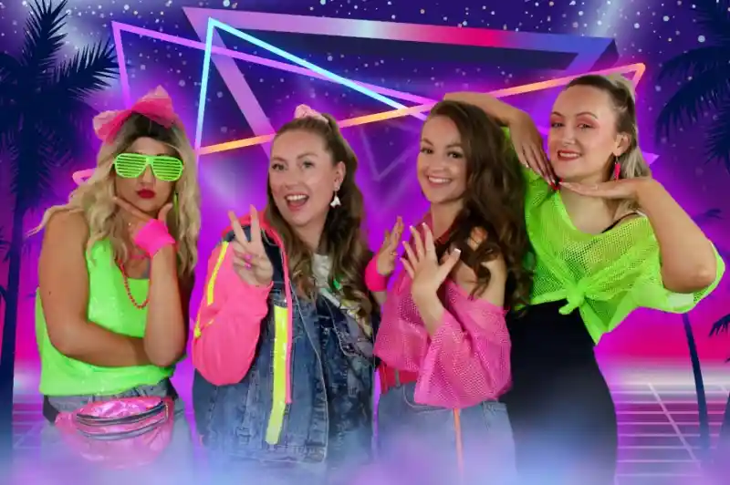 Four women pose together in colorful 1980s-inspired outfits against a vibrant, neon background featuring geometric shapes and palm trees. They display playful expressions and gestures, showcasing their bright accessories and hairstyles.