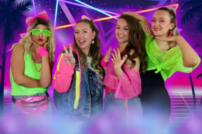 Four women pose together in colorful 1980s-inspired outfits against a vibrant, neon background featuring geometric shapes and palm trees. They display playful expressions and gestures, showcasing their bright accessories and hairstyles.