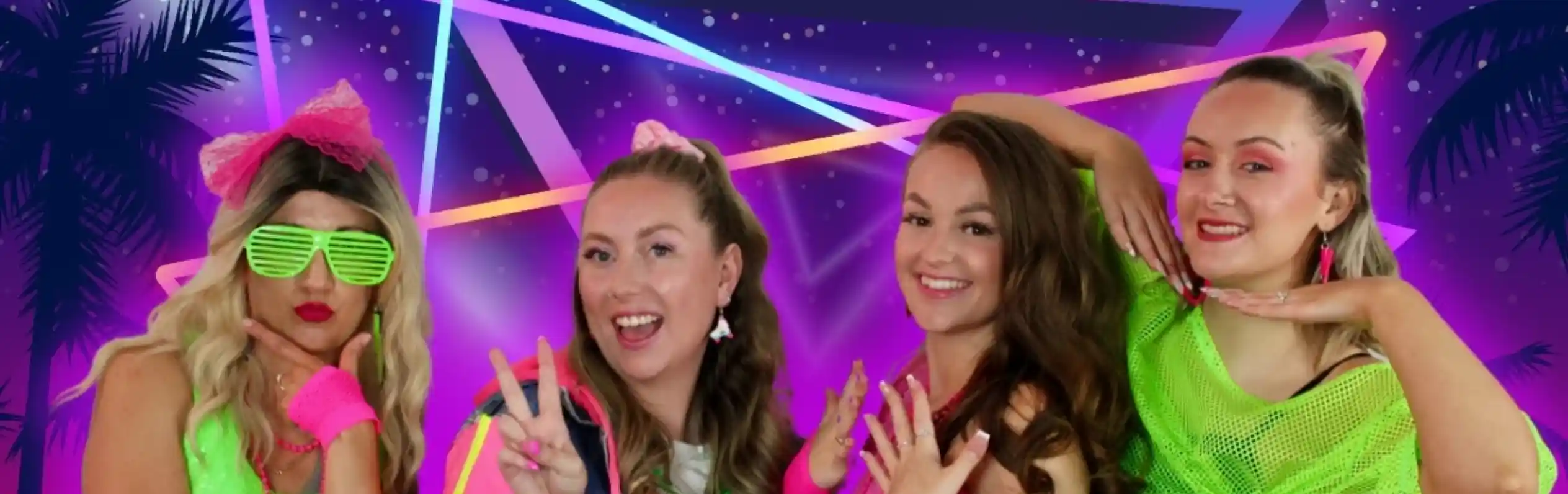 Four women pose together in colorful 1980s-inspired outfits against a vibrant, neon background featuring geometric shapes and palm trees. They display playful expressions and gestures, showcasing their bright accessories and hairstyles.