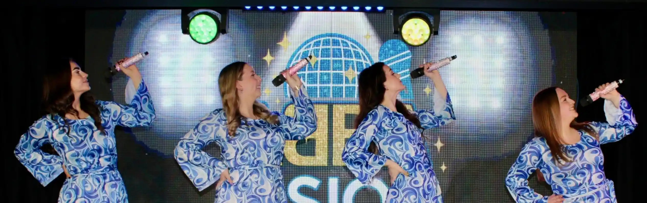 Four female performers in matching blue patterned dresses and white knee-high boots strike dynamic poses while holding microphones. A colorful backdrop features lights and a logo.