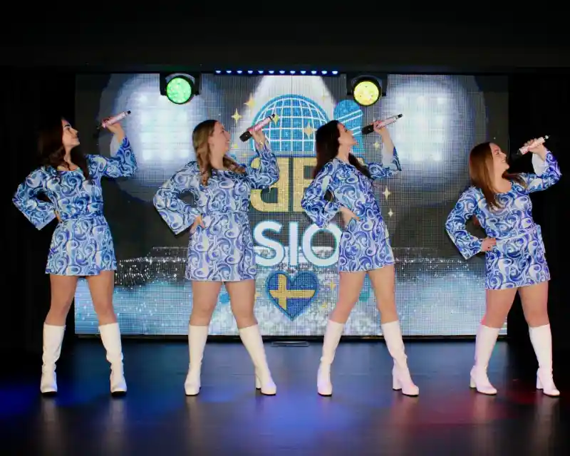 Four female performers in matching blue patterned dresses and white knee-high boots strike dynamic poses while holding microphones. A colorful backdrop features lights and a logo.