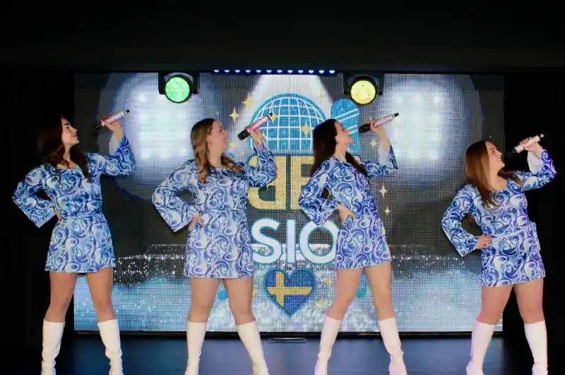 Four female performers in matching blue patterned dresses and white knee-high boots strike dynamic poses while holding microphones. A colorful backdrop features lights and a logo.